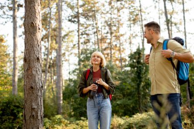 A woman and a man pause in a sunny forest, smiling and chatting. They wear hiking gear, carrying backpacks, surrounded by tall trees and lush greenery, enjoying nature together.