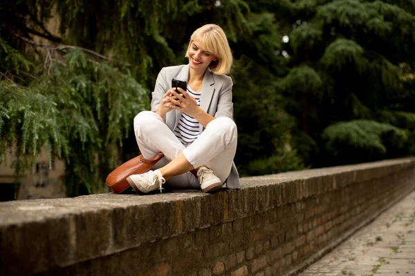 A woman with blonde hair sits cross-legged on a brick wall outside during the daytime. She smiles while looking at a cellphone in her hands with trees in the background.