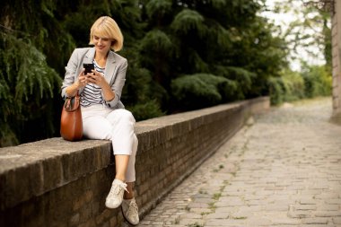 Bir kadın, yeşil bir parkta, akıllı telefonuna odaklanmış, sıradan bir şekilde alçak bir taş duvarda oturuyor. Açık renk bir ceket giyiyor ve rahat görünüyor. Keyifli bir öğleden sonra geçiriyor..