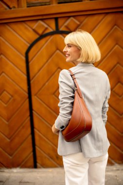 Kısa saçlı genç bir kadın ahşap bir duvarın yanında dururken gülümsüyor. Açık gri bir ceket ve beyaz pantolon giyiyor. Elinde kahverengi bir omuz çantası var..