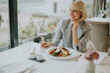 Modern bir restoranda beyaz elbiseli bir masada şık bir kadın oturuyor. Bir gurme yemeğinin ve bir bardak şarabın keyfini çıkarırken gülümsüyor. Büyük pencerelerden içeri güneş ışığı giriyor..