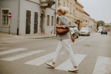 Bir kadın yaya geçidinde yürür. Şehir merkezinde. Açık renkli günlük kıyafetler giyiyor ve şehir ortamında güvenli bir şekilde hareket eden kahverengi bir çanta taşıyor..