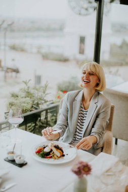 Kısa sarı saçlı, gülümseyen bir kadın şık bir restoranda bir masada oturup lezzetli bir yemeğin tadını çıkarıyor. Güzel rıhtım manzarası yemek deneyimini arttırır..