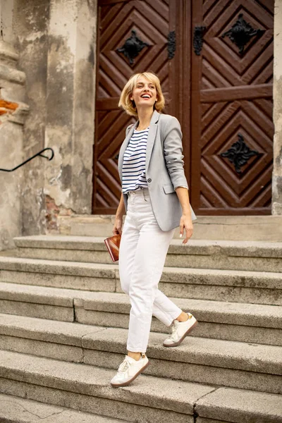 Açık gri ceketli, çizgili bluzlu ve beyaz pantolonlu bir kadın taş basamaklarda mutlu bir şekilde yürüyor. Küçük bir çantası var ve büyük bir ahşap kapının önünde gününün tadını çıkarıyor..