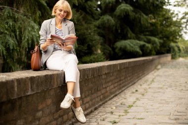 Sarı saçlı bir kadın dışarıdaki alçak tuğlalı bir duvarda oturuyor. O bir kitap okuyor ve gülümsüyor. Yanında bir cüzdanı var. Ağaçlar arka planda..