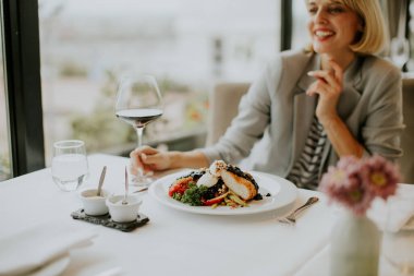 Suya bakan bir restoranda bir masada oturan bir kadın yanında bir bardak kırmızı şarapla gurme yemeğinin tadını çıkarıyor. Anın tadını çıkarırken sıcak bir şekilde gülümsüyor..
