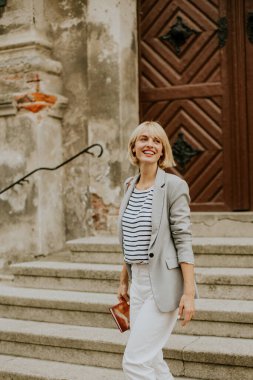 Açık renk ceket ve çizgili bluz giyen genç bir kadın sevinçle taş basamaklardan çıkıyor. Sıradan ama modaya uygun kıyafeti ve parlak gülüşü, tarihi ortamda neşeli bir atmosfer yaratıyor..