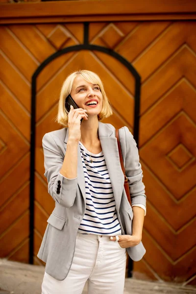 A stylish woman stands confidently in front of an intricately designed wooden door, chatting joyfully on her phone. She wears a trendy outfit, radiating a cheerful vibe on a bright day.