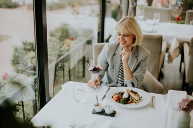 Şık kıyafetli bir kadın şık bir restoranda gurme yemeğinin tadını çıkarırken gülümsüyor. Güneş ışığı büyük pencerelerden geçer ve sıcak bir ortam yaratır..