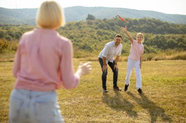 Bir grup arkadaş yeşil tepelerle çevrili güneşli bir arazide frizbi oynuyorlar. Biri frizbiyi atarken, iki insan gülümser ve güler..