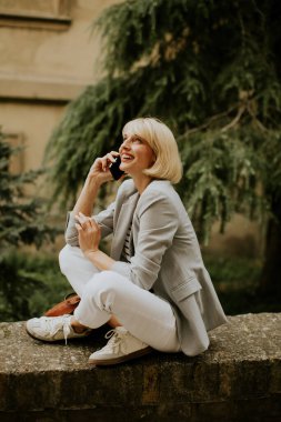Beyaz pantolon giyen sarışın bir kadın ve gri ceketli bir kadın dışarıda taş bir çıkıntıda oturmuş cep telefonuyla konuşuyor ve mutlu bir şekilde gülümsüyor..