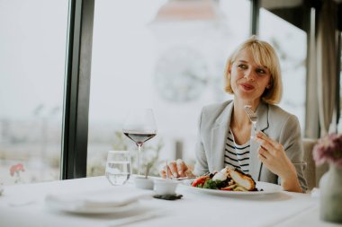 Bir kadın şık bir restoranda masada oturmuş, dikkatlice hazırlanmış yemeğin tadını çıkarıyor. Bir çatal tutuyor ve pencereden şehir manzarasına bakarken gülümsüyor. Bir bardak kırmızı şarapla rahatlıyor..