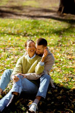 Neşeli çift çimlerin üzerinde birlikte oturur, birlikte güler ve neşeli bir güneş parkında eğlenirler. Renkli sonbahar yapraklarıyla çevrili..