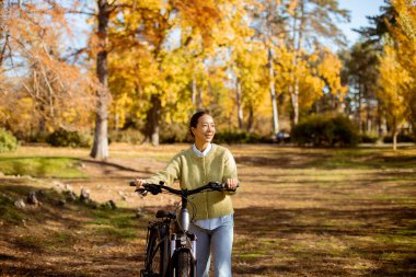 Koreli bir kadın, rengarenk yapraklar ve sıcak güneş ışığıyla çevrili, canlı bir parkta bisikletiyle mutlu bir şekilde duruyor..