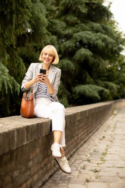 Taş bir duvarda oturan bir kadın, neşeli bir ifadeyle akıllı telefonuna bakıyor. Serin bir bahçede yemyeşil bir yeşil ile çevrili açık renk bir ceket ve beyaz bir pantolon giyiyor..