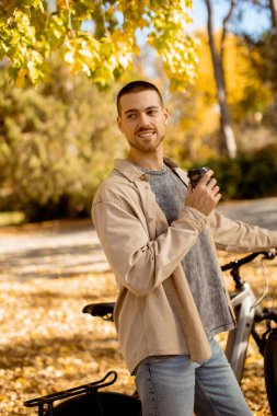 Genç bir adam altın güneş altında güneşlenen renkli sonbahar yapraklarıyla dolu bir parkta bisikletine yaslanırken bir fincandan yudumluyor..