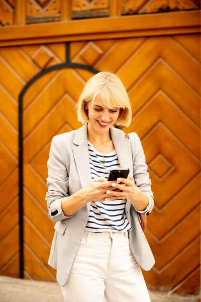 Sarı saçlı ve çizgili tişörtlü bir kadın akıllı telefonuyla güzel bir ahşap duvarın önünde mutlu mesut mesaj atıyor. Sahne onun rahat ve neşeli tavrını yakalar..