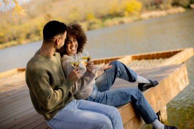 İki kişi ahşap bir rıhtımda oturmuş, gülümsüyor ve elinde köpüklü içecek bardakları tutuyordu. Güneş arkalarında batar, ılık bir parıltı yaratır..