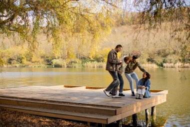 Dört arkadaş, sonbahar renkleriyle çevrili sakin bir gölün kenarındaki ahşap rıhtımda kahkaha ve neşe dolu anları paylaşıyor..