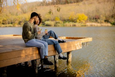 İki insan, sonbahar ağaçlarıyla çevrili sakin bir gölün kıyısındaki ahşap iskelede otururken içkinin keyfini çıkarıyor..