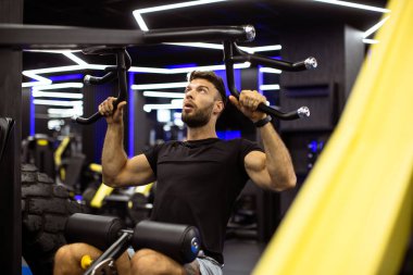 Bir adam spor salonunda bir çekme makinesi kullanıyor. O, barı indirirken odaklanıyor ve kendini yoruyor. Spor salonu çağdaş tasarım özellikleriyle aydınlık..
