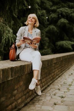 Genç bir kadın gün boyunca bir parkta taş bir duvarda oturuyor. O bir kitap okurken gülümser. O bir ceket ve beyaz pantolon giyiyor ve bir omuz çantası onun yanında dinleniyor..