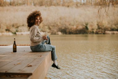 Genç bir kadın sakin bir gölün kıyısında oturur, içkisini yudumlar ve akşam ışığı suyu yansıtırken gülümser..
