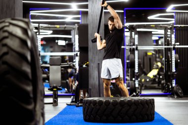 Bir adam enerji dolu bir şekilde çekiçle büyük bir lastiğe vuruyor. Pürüzsüz, modern bir spor salonunda..