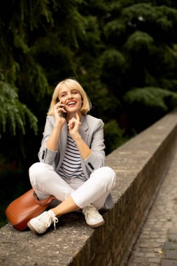 Neşeli bir kadın yeşil bir parkta taş bir duvarda oturmuş, telefonuyla konuşuyor. Çizgili bir gömleğin, beyaz bir pantolonun üzerine açık renk bir ceket giyiyor ve güneşin altında parlak bir şekilde gülümsüyor..