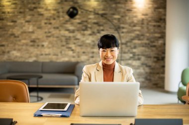 Profesyonel bir kadın dizüstü bilgisayarı açık bir şekilde masasında oturuyor ve kendine güvenerek gülümsüyor. Sıcak, modern ofis taş duvarlar ve şık mobilyalar içeriyor ve iş için hoş bir ortam yaratıyor..