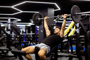 İyi aydınlatılmış bir spor salonunda göğüs presi yapan bir adam. Göğsünün üzerinde çeşitli egzersiz ekipmanlarıyla çevrili halteri kaldırırken konsantrasyonunu gösteriyor. Atmosfer enerjik ve motive edici..