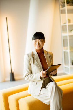 Kendine güvenen iş elbiseli bir kadın sarı bir koltukta oturur, modern, şık bir ofis alanında tablet kullanırken gülümser..