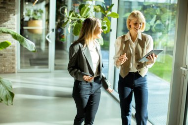 İş kıyafeti giymiş iki kadın, yeşillik ve doğal ışıkla dolu çağdaş bir ofiste yürürken fikirleri paylaşıyor ve ilham verici bir çalışma ortamı yaratıyorlar..