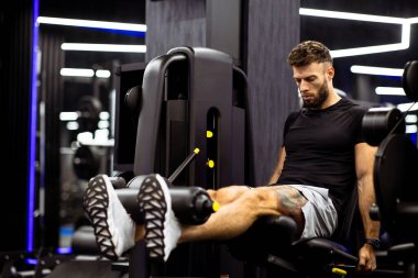 Modern bir spor salonunda bacak uzatma makinesi üzerinde egzersiz yapan bir adam. Bacak dayanıklılığına odaklanıyor. Spor salonu aynalar ve parlak tasarımlarla dolu, enerjik bir atmosfer yaratıyor. Lleg uzantısı