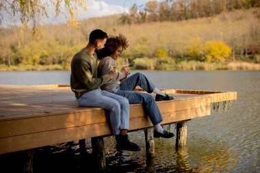 İki arkadaş sakin bir gölün kıyısındaki ahşap iskelede oturur, içkilerini yudumlar ve ılık sonbahar güneşinin altında gülümsemelerini paylaşırlar..