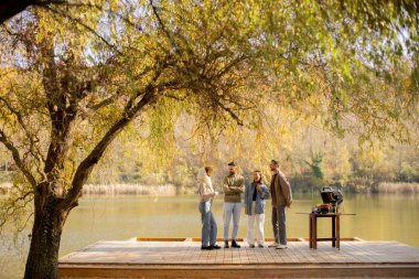 Dört arkadaş, yapraklı bir ağacın altındaki ızgarada yemek hazırlarken sakin bir göl kenarında gülüşlerini ve hikayelerini paylaşıyorlar..