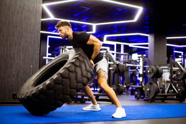 Bir adam şık bir spor salonunda lastik fırlatma egzersizi yapar. Ayar, çeşitli egzersiz ekipmanlarıyla ayrılmış bir fitness alanını gösteren parlak mavi ışıkları içeriyor..