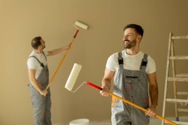 İki adam boya silindirli bir odayı boyamakla meşgul. Tulum giyerler ve duvar rengini değiştirmeye odaklanırlar. Arka planda araçlar ve merdiven görülebilir.
