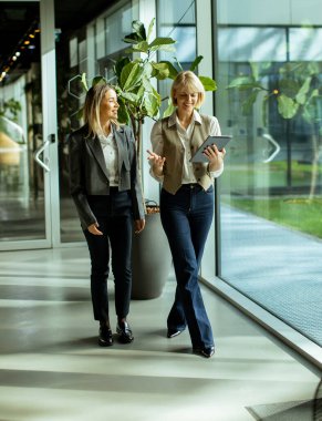 Işıl ışıl yanan bir ofiste, elinde tabletle sohbet eden iki kadın. Yeşilliklerle çevrili şık bir iş kıyafeti giyiyorlar..