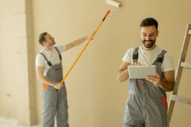 İş tulumlu iki adam iç duvarı yeniden boyuyor. Biri boya uygulamak için silindir kullanırken, diğeri yenilenmiş bir odadaki tablet üzerindeki görevleri kontrol ediyor..