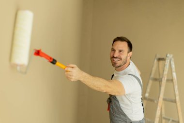 Bir adam açık bej renkli bir silindirle bir duvarı boyarken gülümsüyor. Tulum giyiyor ve merdivenin önünde duruyor. Ev dekorasyonunun keyfini çıkarıyor..