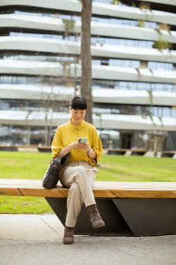 Huzurlu bir şehir parkında, sarı gömlekli bir kadın ahşap bir bankta telefonuna odaklanarak rahatça oturuyor. Binalar ve palmiye ağaçları rahat bir atmosfer yaratır..