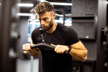 Bir adam akşamları spor salonunda çalışıyor. Bir kablo makinesi kullanıyor, gücünü ve uygunluğunu arttırmak için egzersiz rutinine odaklanıyor..