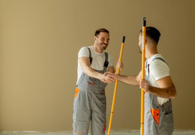 Gri iş tulumlu iki adam temiz, yenilenmiş bir odada gülümseyip el sıkışıyor. Uzun boya rulolarını tutuyorlar, boyama görevlerine birlikte başlamaya hazırlar..