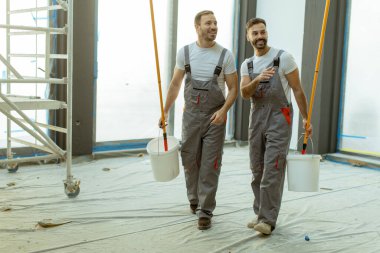 İki gülen işçi ellerinde boya kovası olan bir inşaat alanından geçiyor. Bölgede inşaat malzemeleri var ve iyi aydınlatılmış, bu da aktif yenileme çalışması yapıldığı anlamına geliyor..