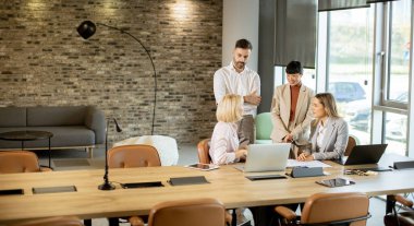Çeşitli bir ekip, uzun tahta bir masanın etrafında bir iş toplantısı yapıyor. Dizüstü bilgisayarlar ve belgeler mevcutken fikirleri tartışıp, ortak bir atmosfer yaratıyorlar..