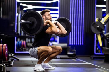 Çağdaş bir spor salonunda bir adam halterle çömeliyor. Enerjik ışıklandırma ve ekipmanlar, dayanıklılık eğitimi ve spor meraklıları için enerjik bir atmosfer yaratır..