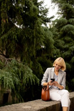 Açık renk elbiseli bir kadın uzun ağaçlarla çevrili taş bir duvarda oturuyor. Akıllı telefonuna odaklanmış, doğada huzurlu bir anın tadını çıkarıyor..