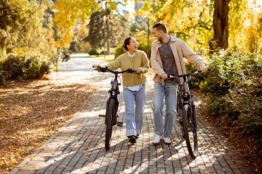 Koreli bir kadın ve beyaz bir adam canlı bir sonbahar parkında yürüyorlar, bisikletlerini tutuyorlar ve renkli ağaçların altında birlikte gülüyorlar..