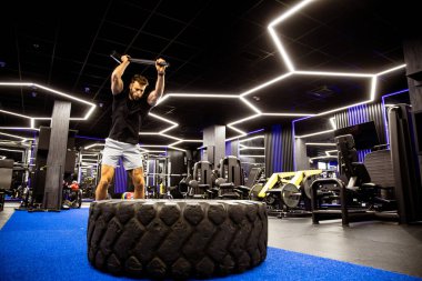 Bir adam şık bir spor salonunda yoğun bir şekilde antrenman yapıyor, büyük bir lastiğe çekiçle vuruyor. Parlak ışıklar ve ekipmanlar antrenman için enerji verici bir atmosfer yaratıyor..